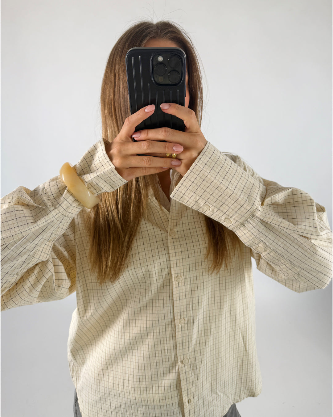En person iført en ternet skjorte tager et billede med en sort smartphone, mens de står i et lyst rum. De har en stor lys armbøjle på venstre håndled.