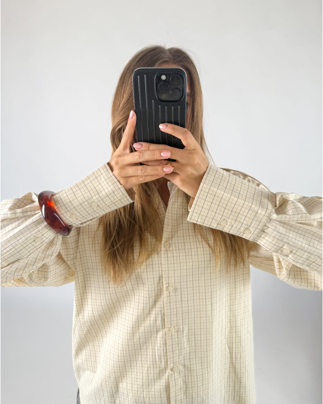 En person holder en mobiltelefon op foran ansigtet, iført en ternet skjorte og et rødt armbånd, i et lyst rum.