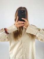 En person holder en mobiltelefon op foran ansigtet, iført en ternet skjorte og et rødt armbånd, i et lyst rum.
