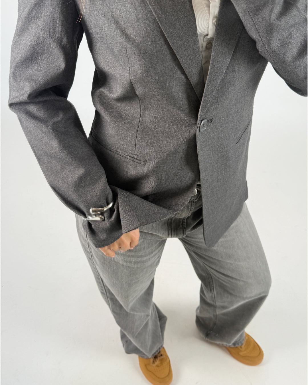 En person iført en grå blazer og vide grå bukser står oprejst, set fra en øvre vinkel. Bagved er der en hvid baggrund, og de har brune sko på.