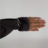 En arm er klædt i en mørkegrå skjorte, hvor ærmet har en skinnende metalring som dekoration. Hånden er strakt ud med lyse, velplejede negle mod en neutral baggrund.