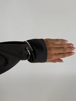 En arm er klædt i en mørkegrå skjorte, hvor ærmet har en skinnende metalring som dekoration. Hånden er strakt ud med lyse, velplejede negle mod en neutral baggrund.