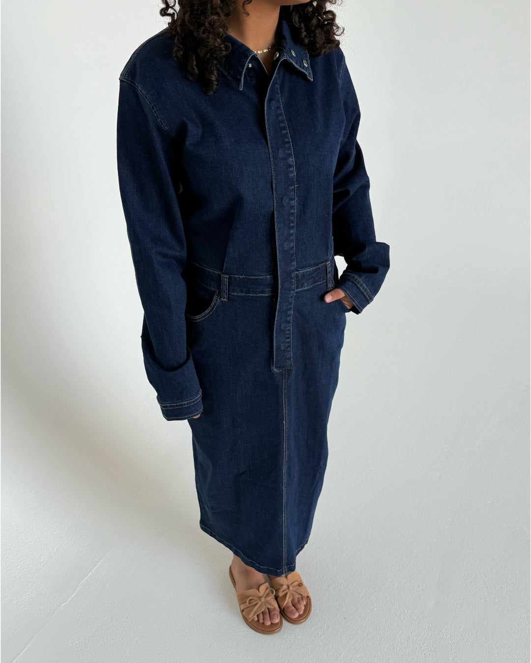En person iført en lang, mørk denimkjole står i et lyst rum. Kjolen har knapper og lange ærmer, og personen bærer sandaler med sløjfer.