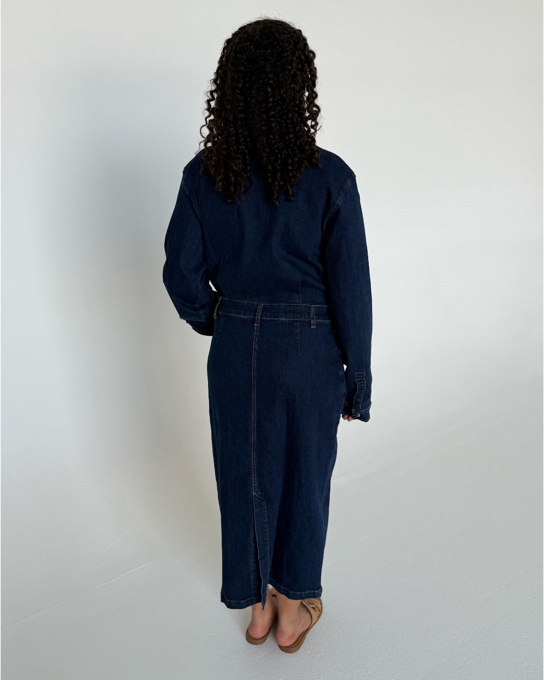 Person iført lang, mørk denimkjole, står med ryggen til, på en lys baggrund. Personen har krøllet hår og bærer sandaler.