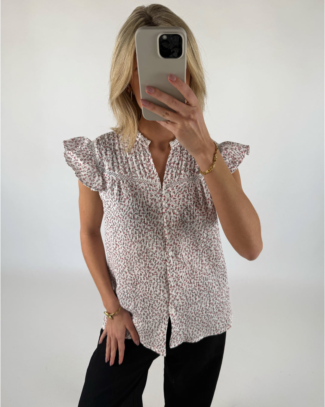 Kvinde iført blomstret bluse tager selfie med mobiltelefon i et lyst rum. Hun har sort tøj på og bærer et guldarmbånd.