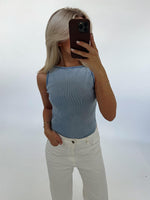 En person iført en lyseblå top og hvide bukser tager et spejlbillede med en mobiltelefon i en minimalistisk, lys baggrund.