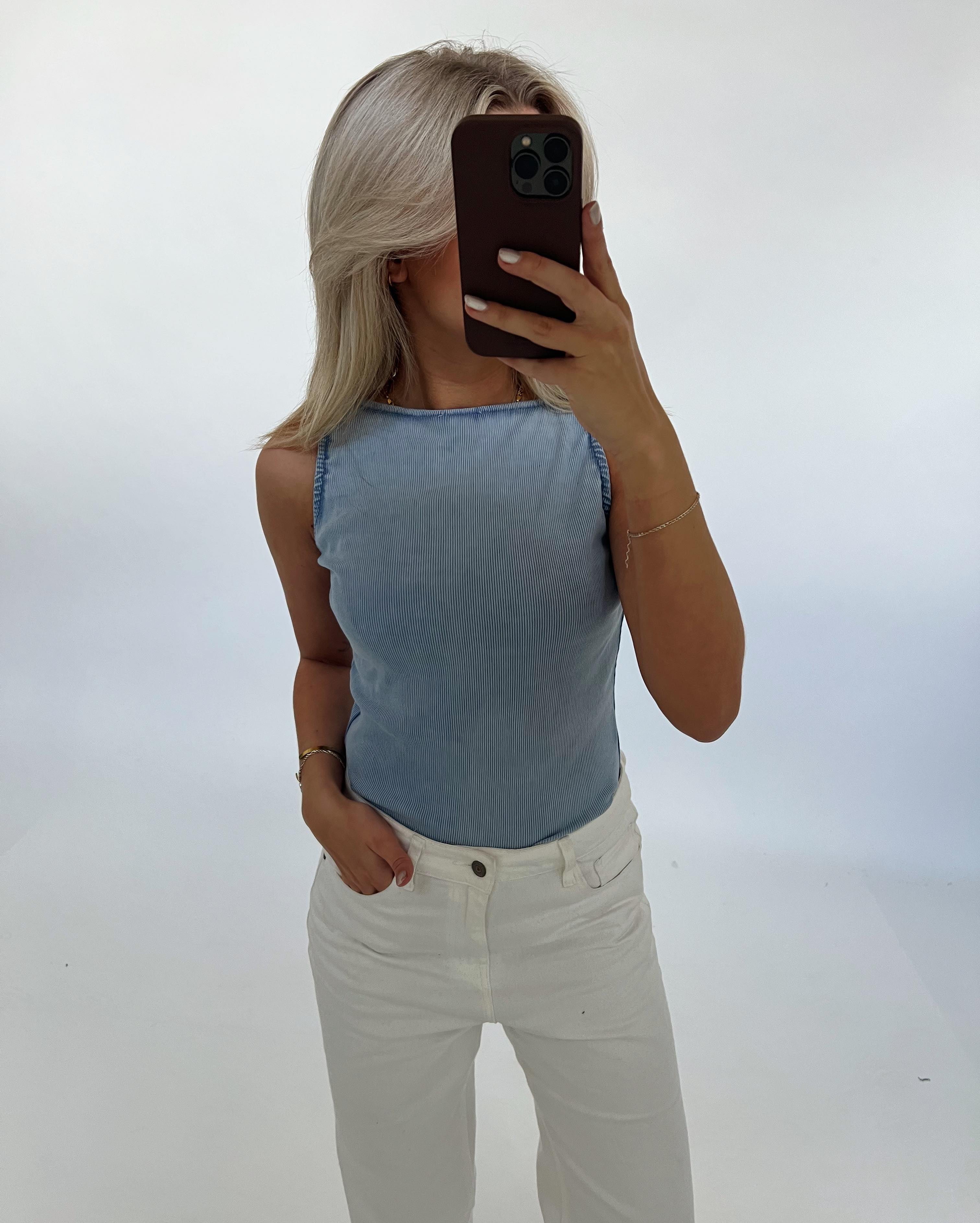 En person iført en lyseblå top og hvide bukser tager et spejlbillede med en mobiltelefon i en minimalistisk, lys baggrund.