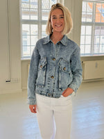 En person iført denimjakke smiler i et lyst rum med hvide vægge og store vinduer, der lukker naturligt lys ind. Personen har hånden i lommen.