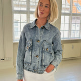 En person iført en lyseblå denimjakke står og laver en ansigtsudtryk i et lyst, rummeligt værelse med store vinduer og hvidmalede gulve.