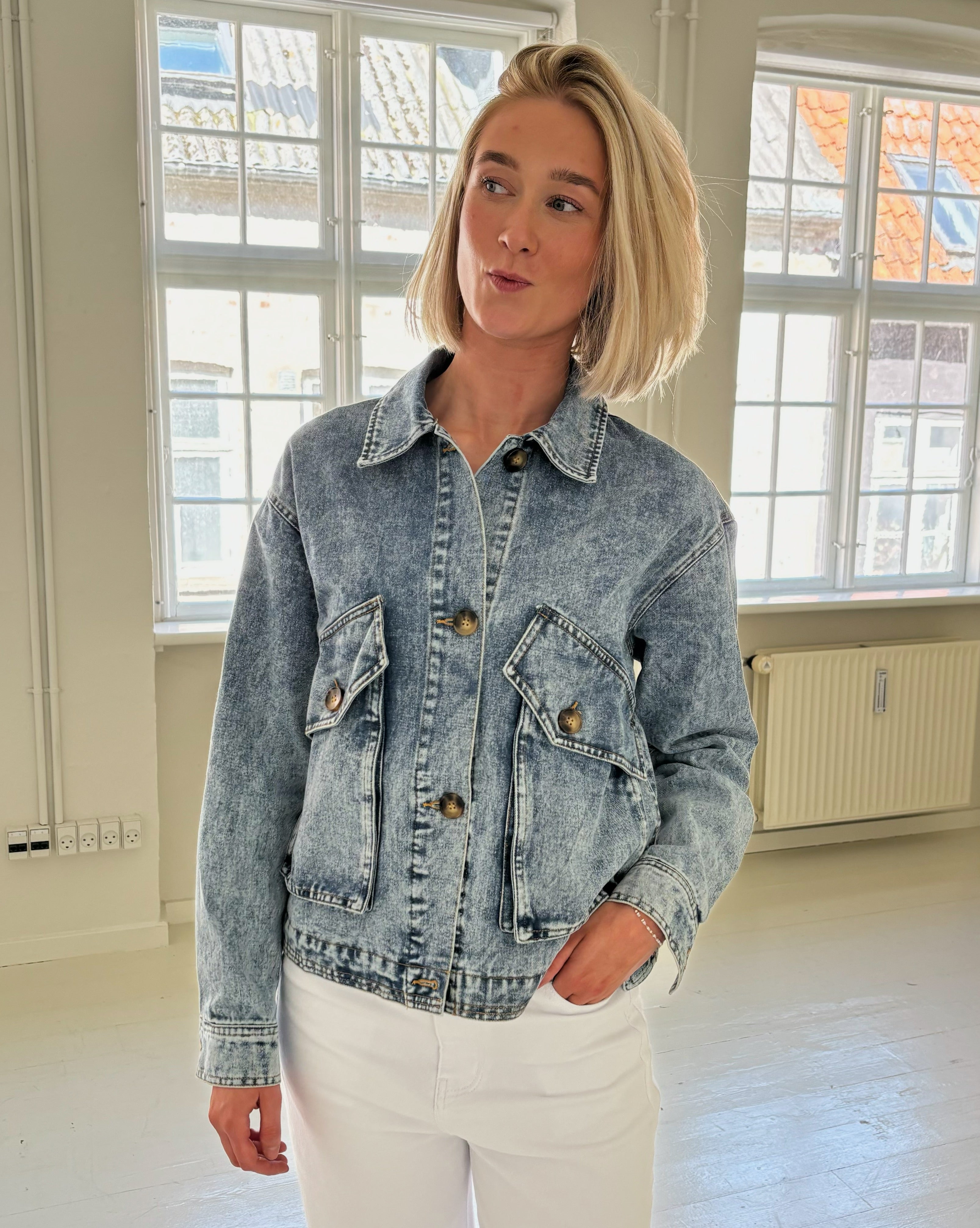 En person iført en lyseblå denimjakke står og laver en ansigtsudtryk i et lyst, rummeligt værelse med store vinduer og hvidmalede gulve.