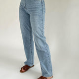 Lyseblå jeans bæres af en person, stående oprejst, iført brune sandaler. Baggrunden er enkel og ensfarvet, hvilket fremhæver tøjets detaljer.