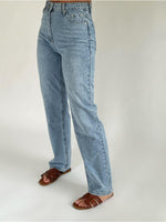 Lyseblå jeans bæres af en person, stående oprejst, iført brune sandaler. Baggrunden er enkel og ensfarvet, hvilket fremhæver tøjets detaljer.