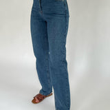 Blå jeans bæres af en person, der står oprejst med brune sandaler. Baggrunden er en ensfarvet hvid væg.