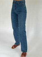 Blå jeans bæres af en person, der står oprejst med brune sandaler. Baggrunden er en ensfarvet hvid væg.