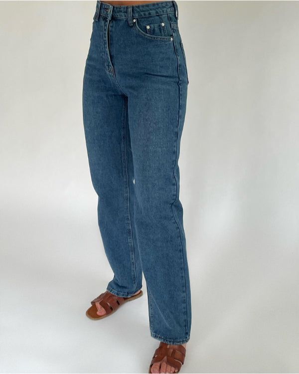Blå jeans bæres af en person, der står oprejst med brune sandaler. Baggrunden er en ensfarvet hvid væg.