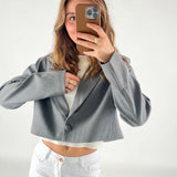 En person iført en grå cropped jakke tager en selfie med en smartphone. Personen har hvidt tøj på under jakken og ringen på venstre hånd. Baggrunden er neutral.