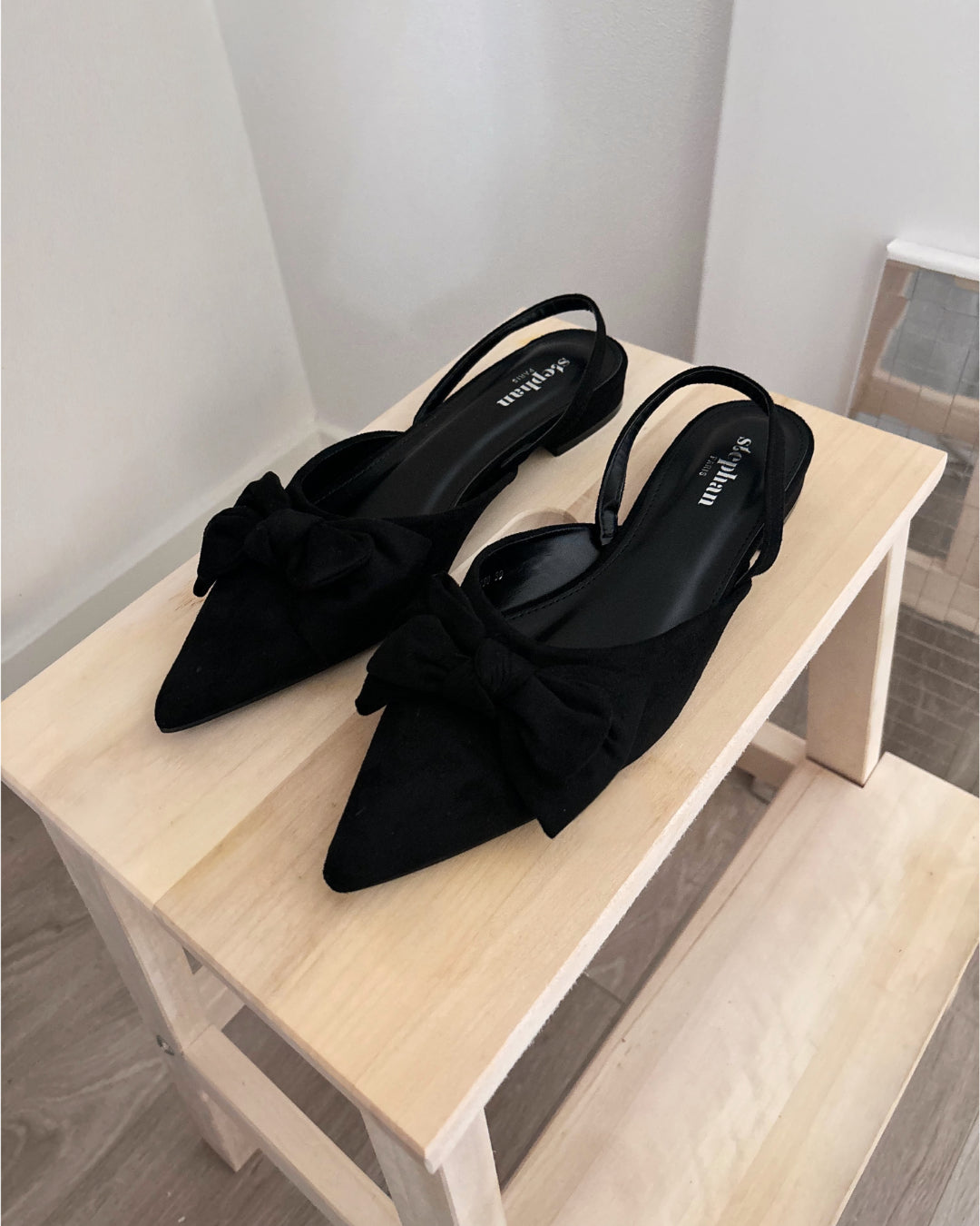 Sort slingback-sko med sløjfer placeret stilfuldt på en lys træhylde i et minimalistisk rum. Skoene har teksten "soft mix" på sålerne.