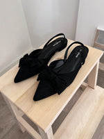 Sort slingback-sko med sløjfer placeret stilfuldt på en lys træhylde i et minimalistisk rum. Skoene har teksten "soft mix" på sålerne.
