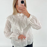 En person iført en hvid bluse med blonder tager et spejlbillede med en lyserød telefon, mod en ensfarvet, lys baggrund.
