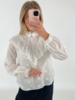 En person iført en hvid bluse med blonder tager et spejlbillede med en lyserød telefon, mod en ensfarvet, lys baggrund.