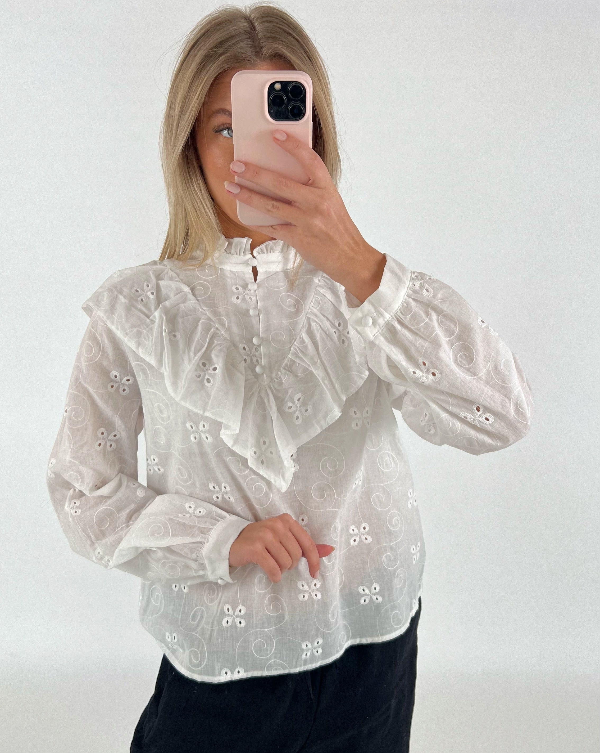 En person iført en hvid bluse med blonder tager et spejlbillede med en lyserød telefon, mod en ensfarvet, lys baggrund.