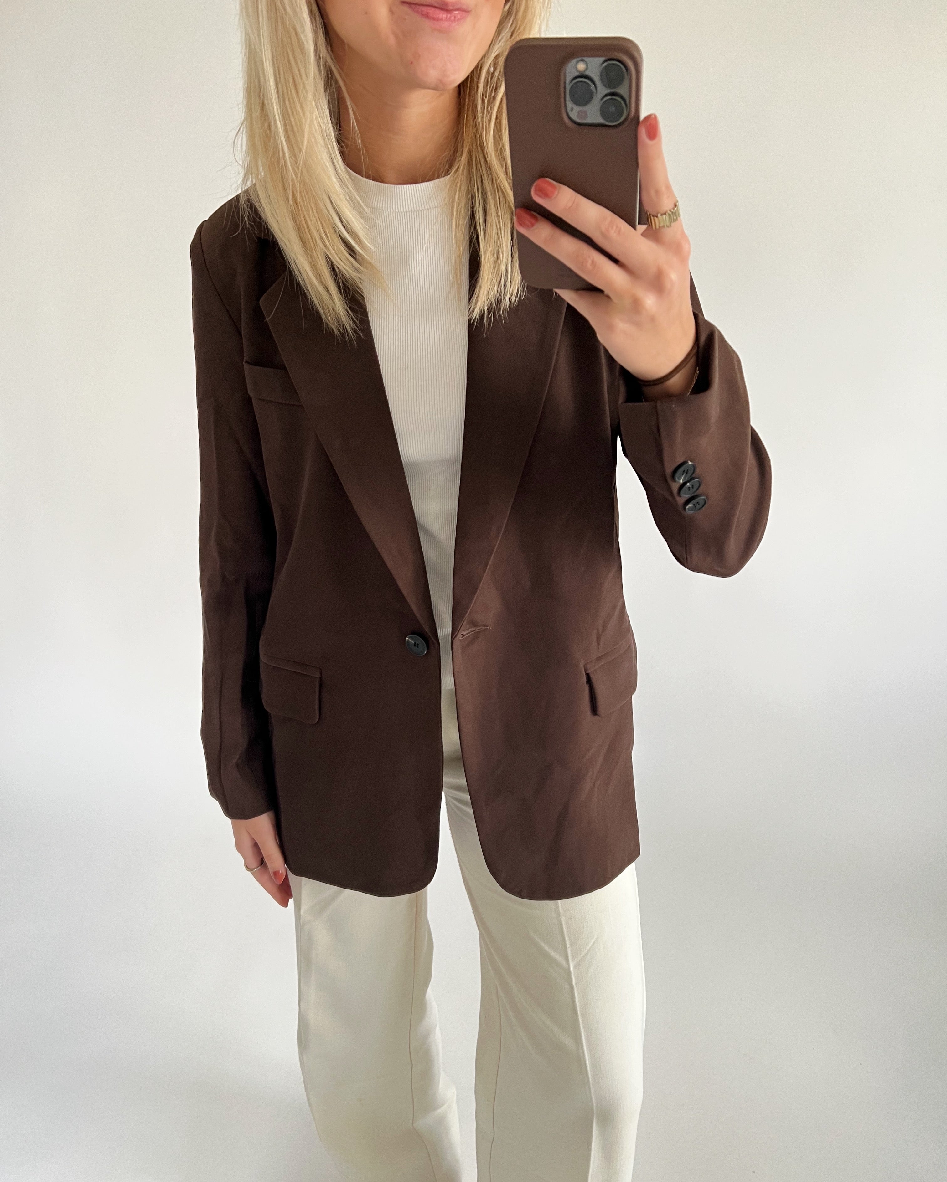 En person iført brun blazer og hvid skjorte tager et selfie med en smartphone. Personen har lyst hår og bærer cremefarvede bukser i et neutralt oplyst rum.
