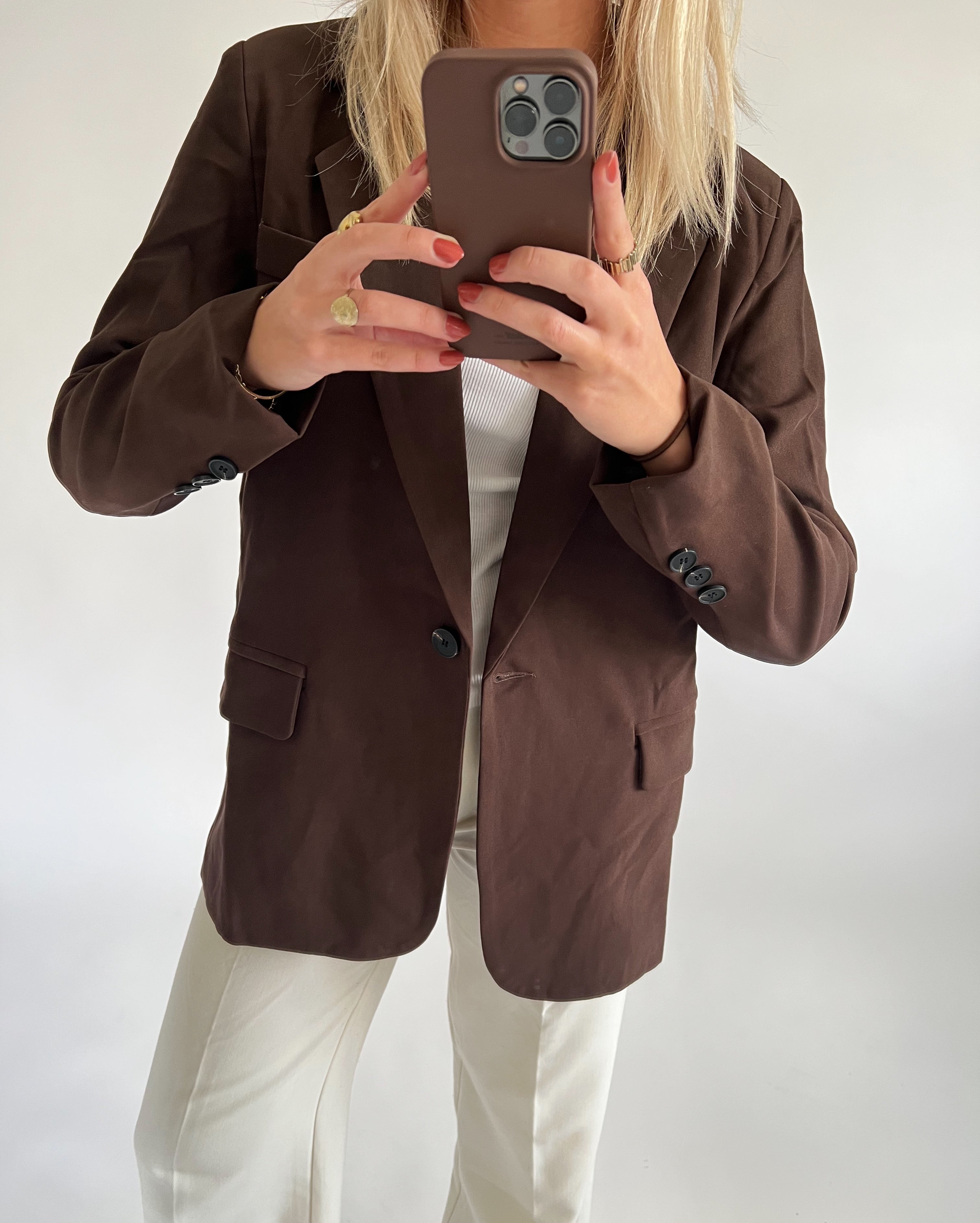 En person iført en brun jakke og hvide bukser tager et spejlbillede med en mobiltelefon, omgivet af en neutral, lys baggrund.