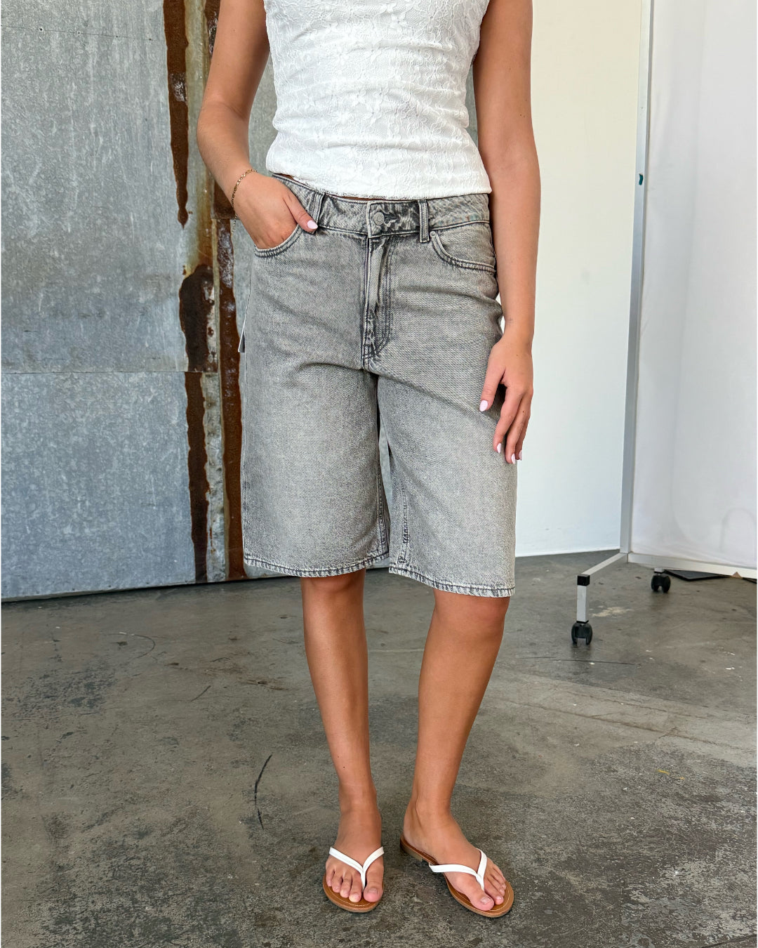 Person iført en hvid blondetop og grå denimshorts står afslappet med en hånd i lommen. Bagved flader med gråtoner og en hvid skillevæg på hjul.