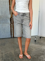 Person iført en hvid blondetop og grå denimshorts står afslappet med en hånd i lommen. Bagved flader med gråtoner og en hvid skillevæg på hjul.