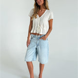 Kvinde står afslappet iført hvid strikket top og lys denim knælange shorts. Hun har sandaler på, hænderne i lommerne og befinder sig i et lyst, minimalistisk rum.