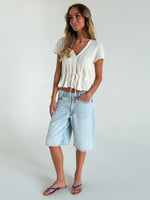 Kvinde står afslappet iført hvid strikket top og lys denim knælange shorts. Hun har sandaler på, hænderne i lommerne og befinder sig i et lyst, minimalistisk rum.