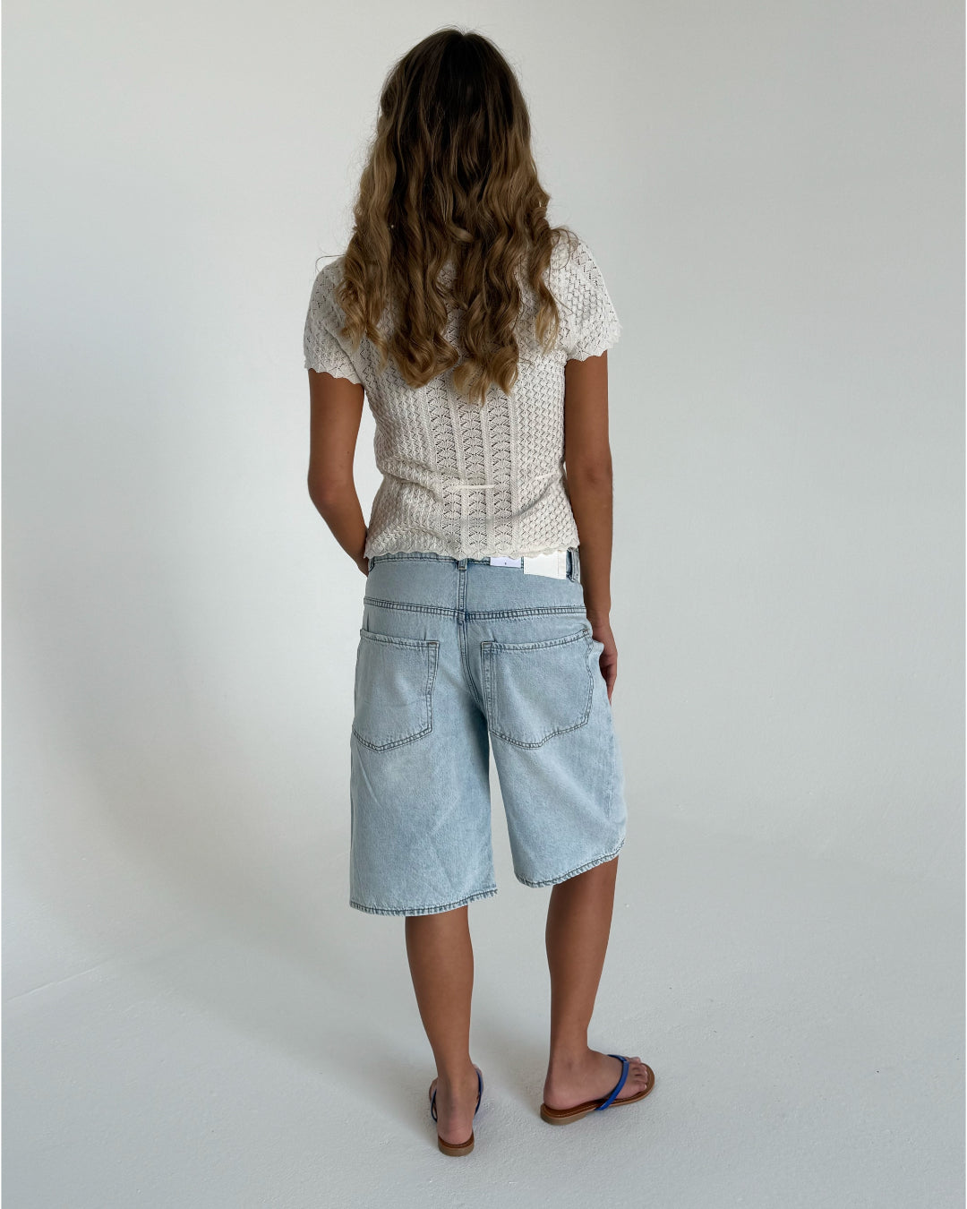En person står med ryggen til, iført en hvid, strikket bluse og lyse denimshorts i en minimalistisk, lys indendørs indstilling. Håret er langt og bølget.