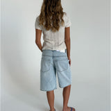 En person står med ryggen til, iført en hvid, strikket bluse og lyse denimshorts i en minimalistisk, lys indendørs indstilling. Håret er langt og bølget.