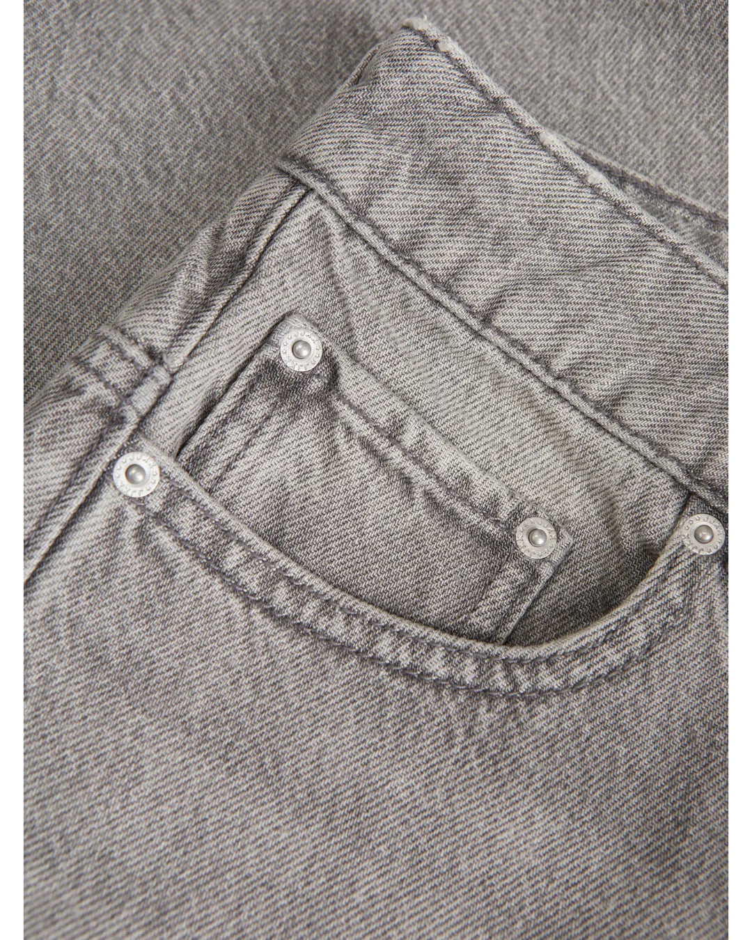 Grå jeanslomme med synlige sømme og metalknapper, placeret mod en baggrund af denimstof i samme farve og tekstur.