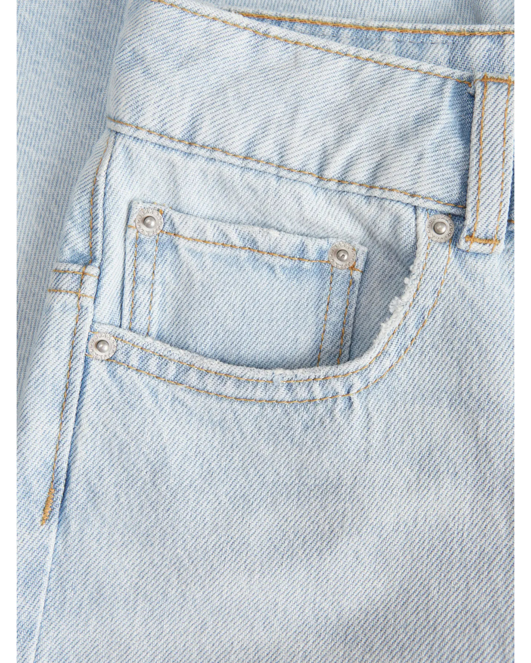 Lyseblå denimstof med synlige lommer og sølvfarvede nitter. Stitching er i kontrasterende gult tråd, og en bæltestrop er synlig. Stoffet har en let struktureret overflade.