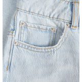 Lyseblå denimstof med synlige lommer og sølvfarvede nitter. Stitching er i kontrasterende gult tråd, og en bæltestrop er synlig. Stoffet har en let struktureret overflade.