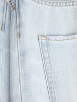 Stoffet er lys denim med gul syning, viser en synlig lomme og en metalknap i øverste venstre hjørne, i en tæt plan flade.