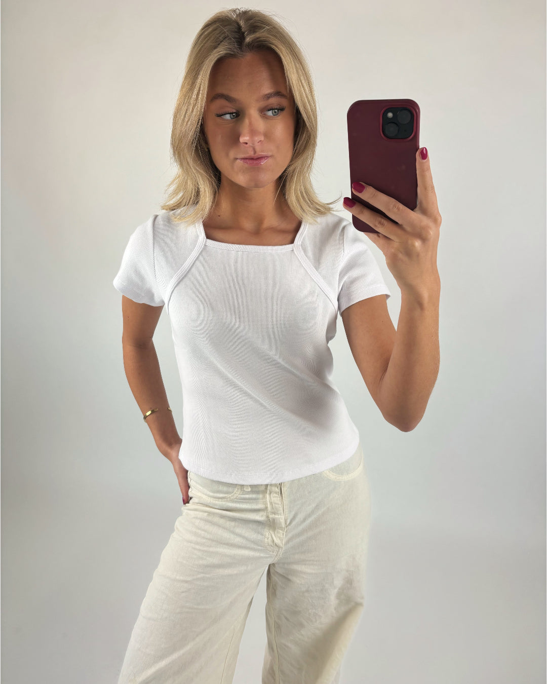 Person tager selfie med smartphone. Personen bærer hvid t-shirt og beige bukser i en minimalistisk, lys baggrund.