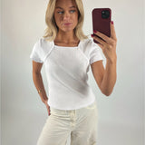 Person tager selfie med smartphone. Personen bærer hvid t-shirt og beige bukser i en minimalistisk, lys baggrund.