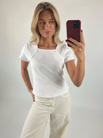 Person tager selfie med smartphone. Personen bærer hvid t-shirt og beige bukser i en minimalistisk, lys baggrund.