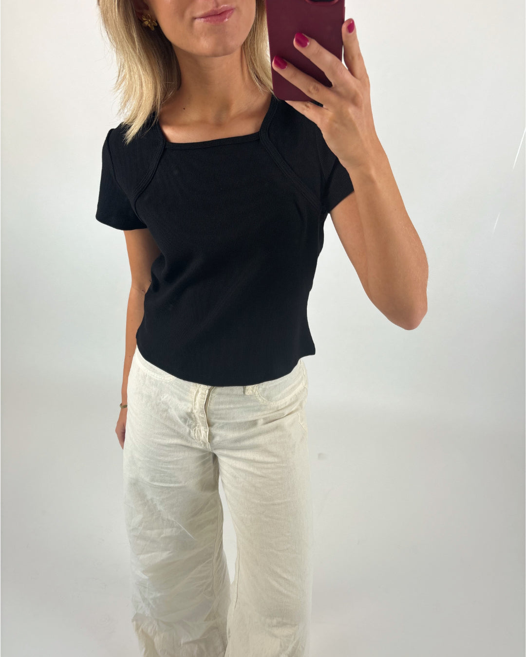 Kvinde iført sort bluse tager selfie med en rød telefon. Hun står i lyse, vide bukser i et lyst rum. Hun har pink neglelak og gyldent blondt hår.
