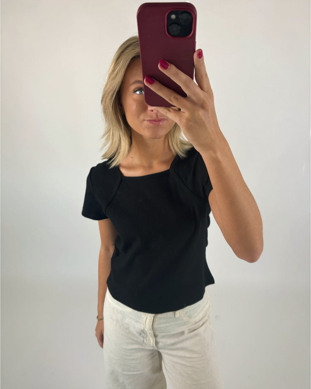 En person holder en rød telefon foran ansigtet, iført sort bluse og hvide bukser, stående i et hvidt, minimalistisk rum.