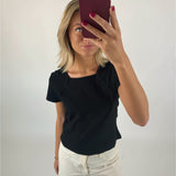 En person holder en rød telefon foran ansigtet, iført sort bluse og hvide bukser, stående i et hvidt, minimalistisk rum.