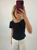 En person holder en rød telefon foran ansigtet, iført sort bluse og hvide bukser, stående i et hvidt, minimalistisk rum.