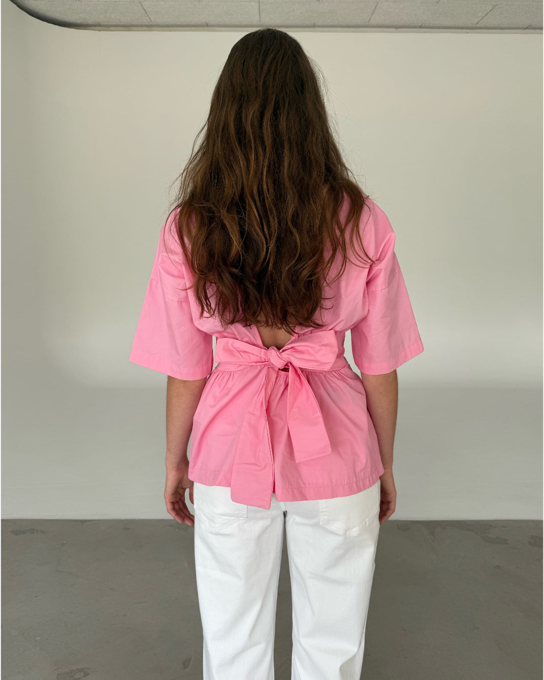 Modelfoto af pige som bærer en lyserød ESTRELLA bluse med bindebånd på ryggen, samt hvide jeans. Alt er fra BySofieSønderby