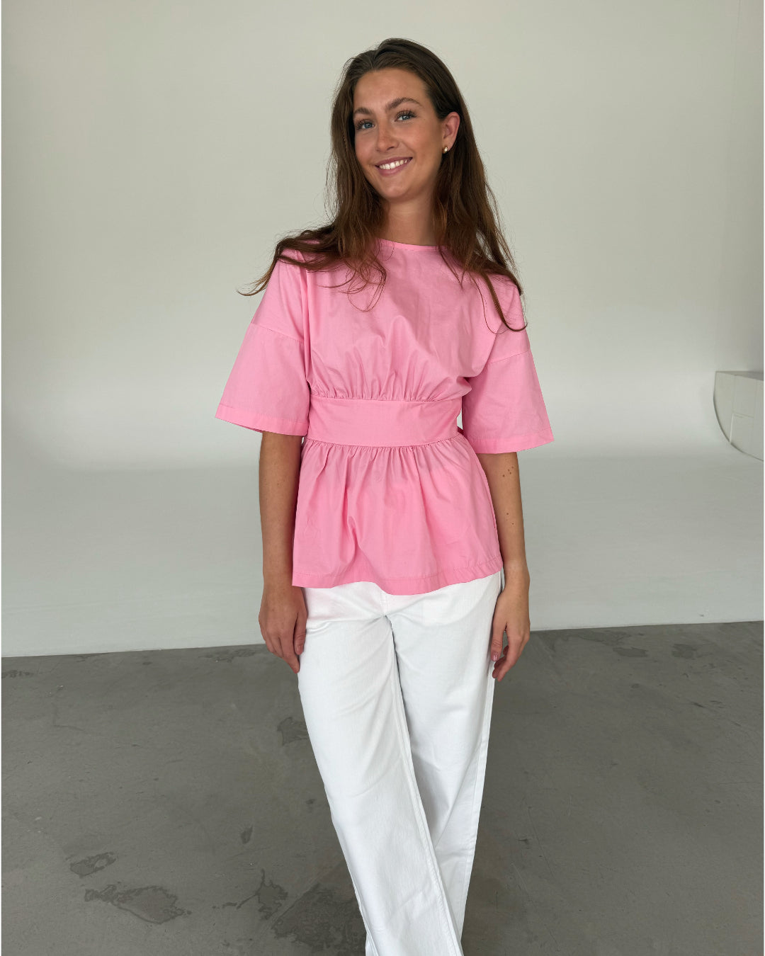 Modelfoto af pige som bærer en lyserød ESTRELLA bluse med bindebånd på ryggen, samt hvide jeans. Alt er fra BySofieSønderby