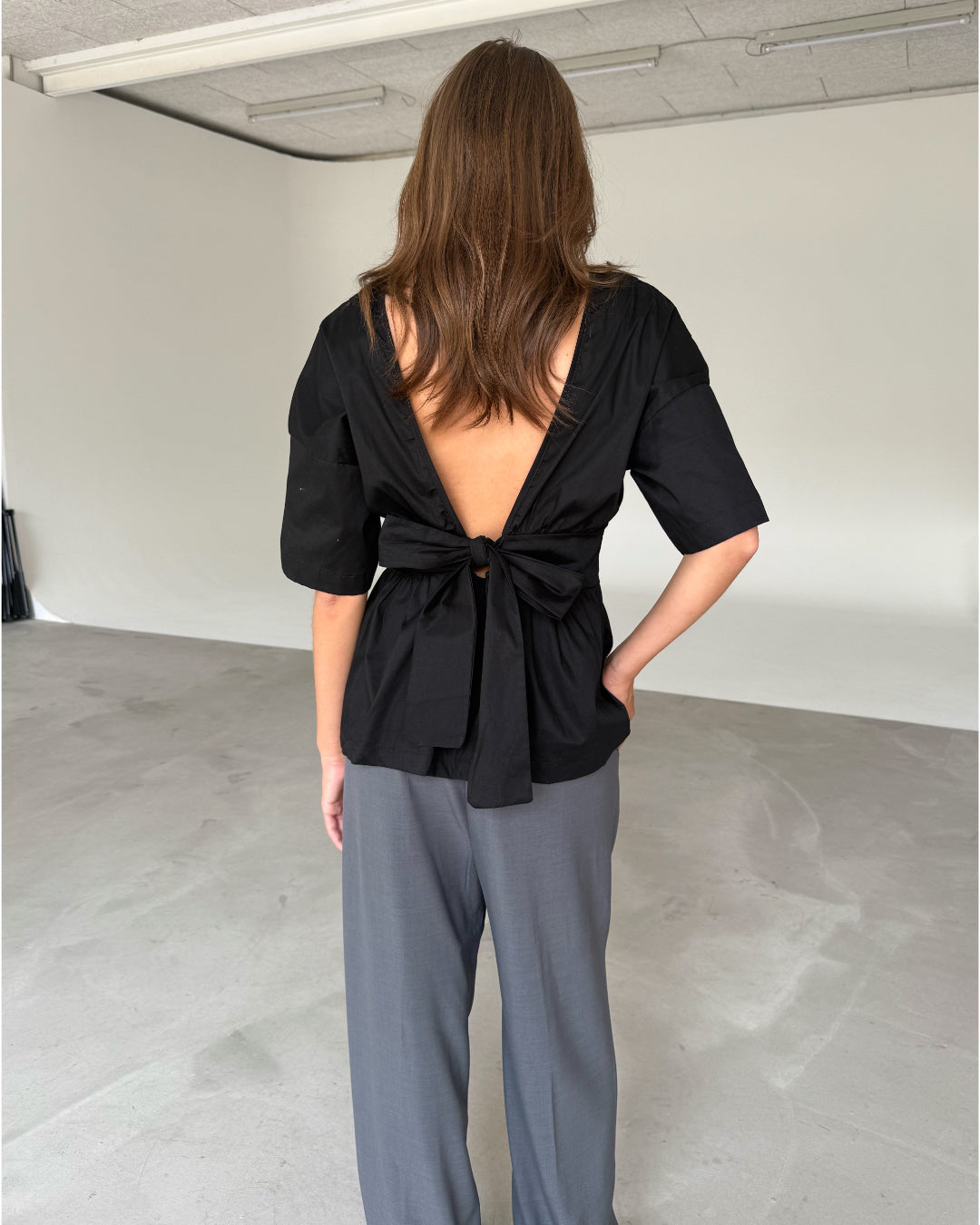 Modelfoto af pige som bærer en sort ESTRELLA bluse med bindebånd på ryggen, samt grå kontorbukser. Set bagfra. Alt er fra BySofieSønderby