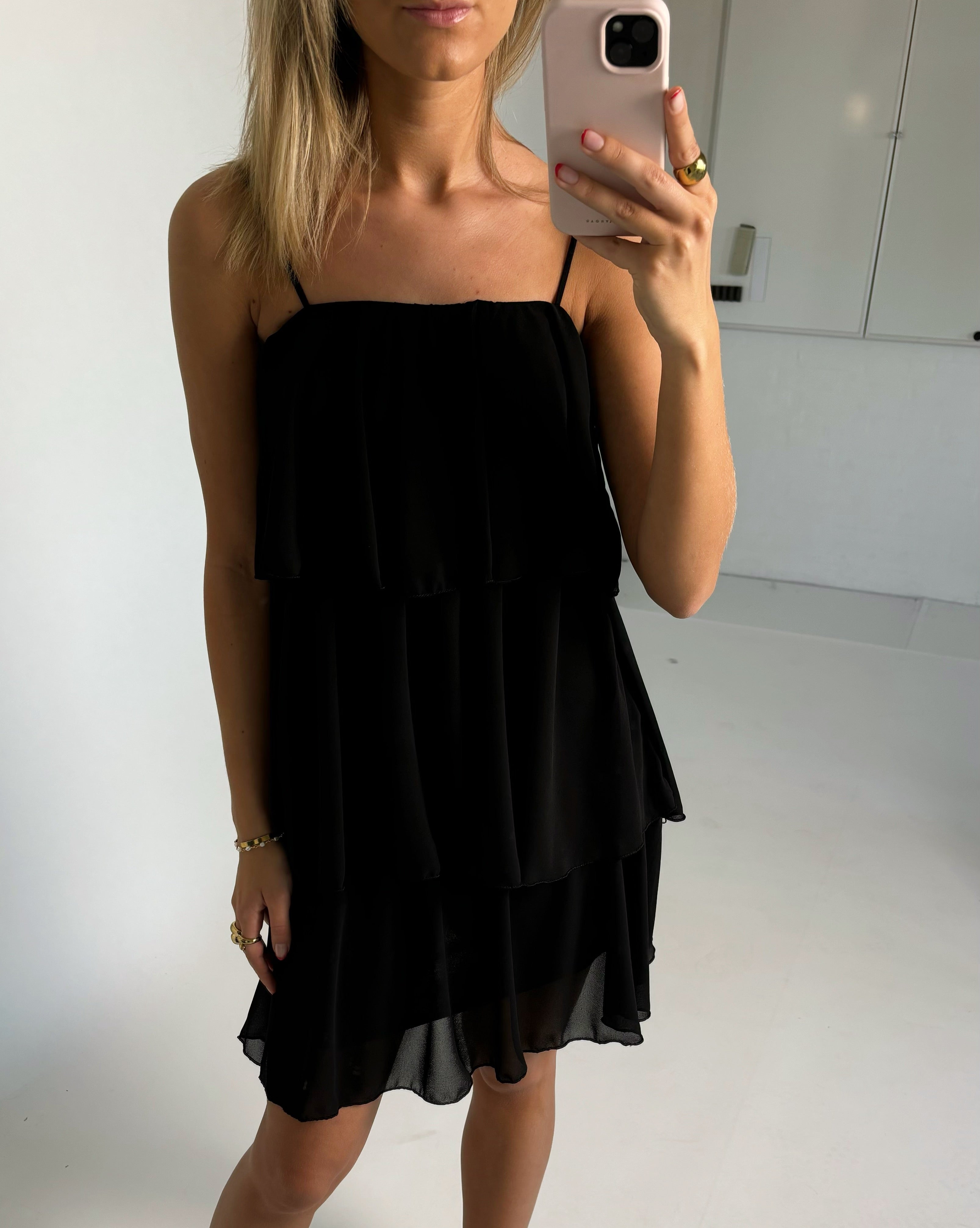 En person iført en sort kjole tager en selfie med en smartphone. Baggrunden er et lyst rum med hvide vægge og skabe.