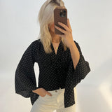 En person tager et spejlbillede iført en sort, prikket bluse og hvide bukser. Personen dækker ansigtet med en mobiltelefon og står i et lyst rum.