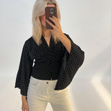 Person tager selfie, iført sort prikket skjorte og hvide bukser, står foran en lys baggrund.